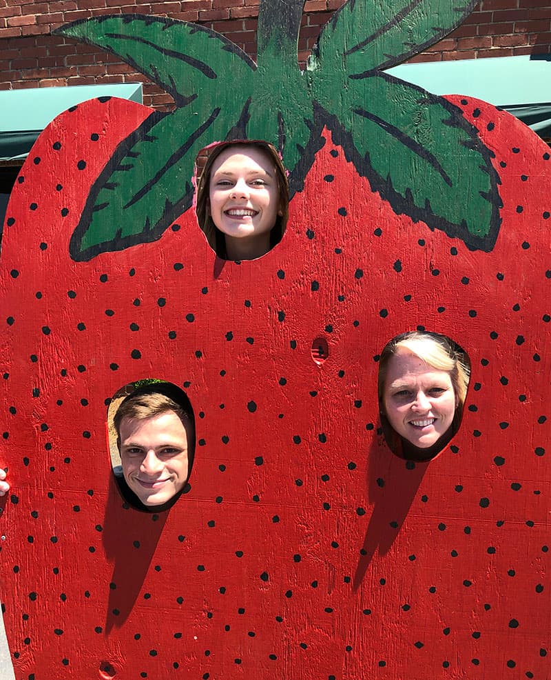 Three people with their faces showing through a wooden strawberry photo prop.